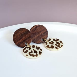 Puede incluir: Pendientes con diseño de madera y cerámica. Los pendientes tienen un disco de madera marrón oscuro y un disco color crema con un estampado de animal marrón. Los pendientes están conectados con aros dorados. Un accesorio elegante.