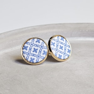 May include: A pair of round stud earrings with a gold-tone border. The earrings feature a white background with a blue floral pattern. The earrings are displayed on a light grey surface.