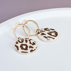 Könnte beinhalten: Ein Paar goldene Creolen mit einem weißen und braunen Leopardenmuster-Anhänger, der an jeder Creole hängt.