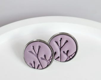 May include: Two round stud earrings with a lavender center and a silver-colored metal frame. The lavender centers feature a dark purple, stylized botanical design. The earrings are displayed on a white surface.