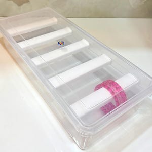 K&ouml;nnte beinhalten: Eine transparente Armreifbox aus Kunststoff mit transparentem Deckel. Die Box hat wei&szlig;e Trennw&auml;nde und enth&auml;lt einen Stapel rosa Armreifen. Die Box dient zur Aufbewahrung und Organisation von Schmuck.