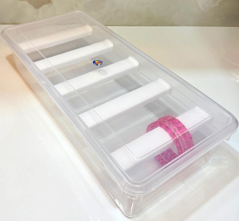 K&ouml;nnte beinhalten: Eine transparente Armreifbox aus Kunststoff mit transparentem Deckel. Die Box hat wei&szlig;e Trennw&auml;nde und enth&auml;lt einen Stapel rosa Armreifen. Die Box dient zur Aufbewahrung und Organisation von Schmuck.
