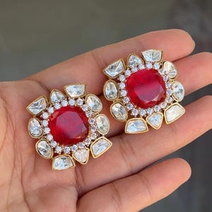 May include: A pair of gold-coloured stud earrings. Each earring has a large, round, deep red gemstone at the centre, encircled by smaller, clear, and irregularly shaped stones. The earrings are designed in a floral pattern.