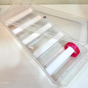 May include: A clear plastic storage box with a hinged lid. Inside are white dividers and a pink and gold bracelet. The box is designed to organize and protect jewelry or small items.