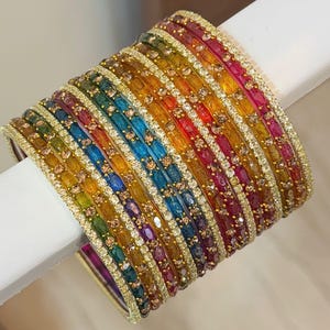 Könnte beinhalten: Ein Stapel bunter Armreifen. Die Armreifen sind in einem Regenbogen aus Farben angeordnet, darunter Rot, Orange, Gelb, Grün, Blau und Lila. Jeder Armreif ist mit kleinen, funkelnden Verzierungen und goldfarbenen Verzierungen versehen.