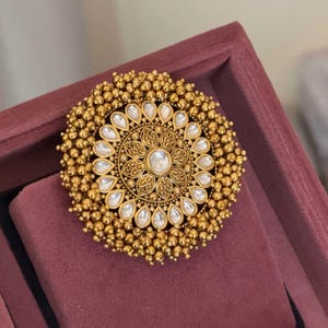 Puede incluir: Un anillo circular de tono dorado que presenta una piedra redonda central rodeada de intrincados trabajos en metal floral. Un anillo de piedras blancas en forma de lágrima rodea el centro, enmarcado por un borde exterior de pequeñas esferas doradas texturizadas.