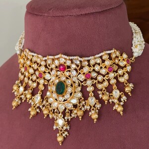 May include: A gold-toned choker necklace featuring an intricate floral design set with clear stones, small pink accents, and a central oval green stone. The piece is embellished with dangling gold beads and small white seed pearls along the edges.