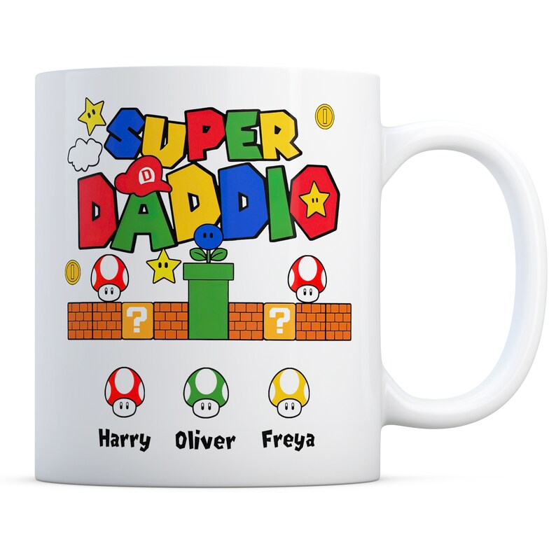 May include: White ceramic mug with a colorful Super Daddi design featuring a Mario-themed graphic with a red, blue, green, and yellow color scheme. The mug has the text "Super Daddi" with a star and a coin. The mug also has three mushrooms with the names Harry, Oliver, and Freya.