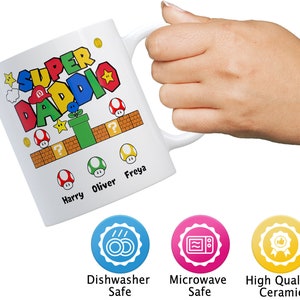 May include: White ceramic mug with a colorful Super Daddie design featuring a video game theme. The mug has the names Harry, Oliver, and Freya printed on it. The mug is dishwasher and microwave safe.