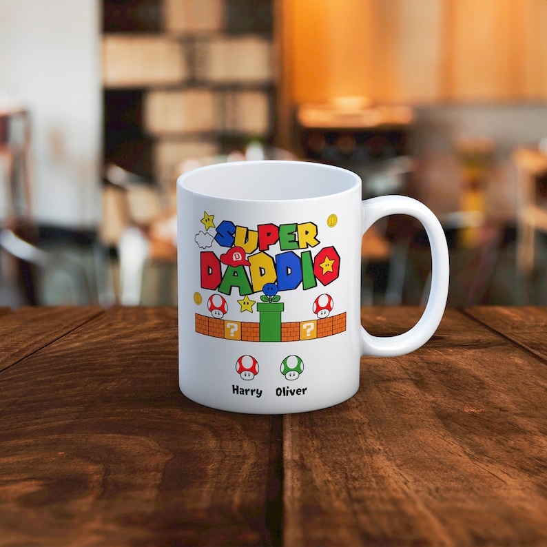 May include: White ceramic mug with a colorful Super Daddi design featuring a video game character. The mug has a green, red, and yellow color scheme and includes the names Harry and Oliver.