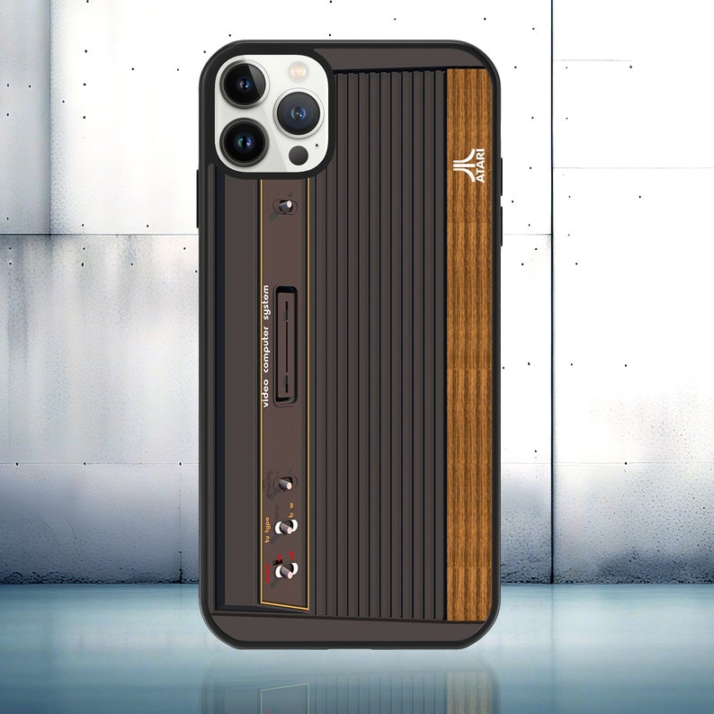May include: A black phone case with a wood-grain design and a retro Atari video computer system graphic. The case has a white camera lens area with three lenses.