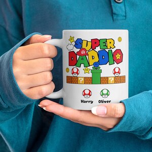 May include: White ceramic mug with a colorful Super Daddi design featuring a video game character and the names Harry and Oliver.