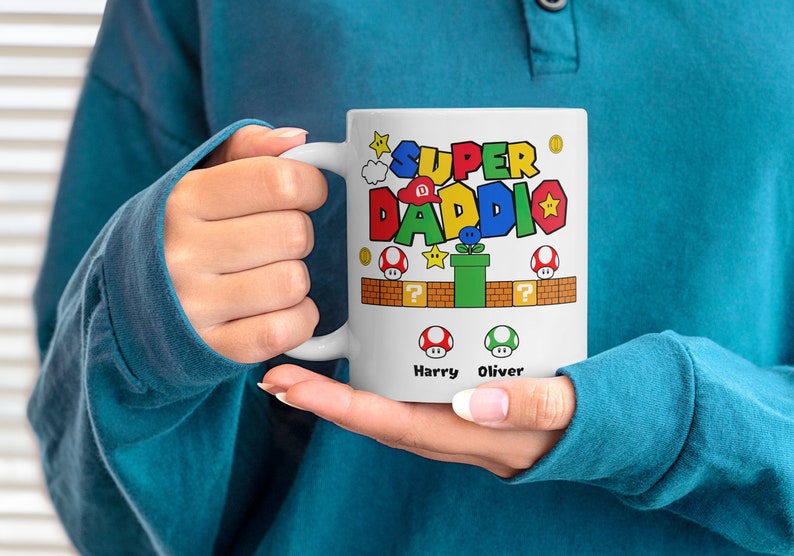 May include: White ceramic mug with a colorful Super Daddi design featuring a video game character and the names Harry and Oliver.