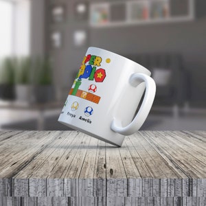 May include: White ceramic mug with a colorful Super Mario Bros. design. The mug features the text "Super Mario" and a yellow star. There are also images of a green pipe, a question mark block, and three mushrooms. The text "Freya" and "Amelia" are written below the mushrooms.