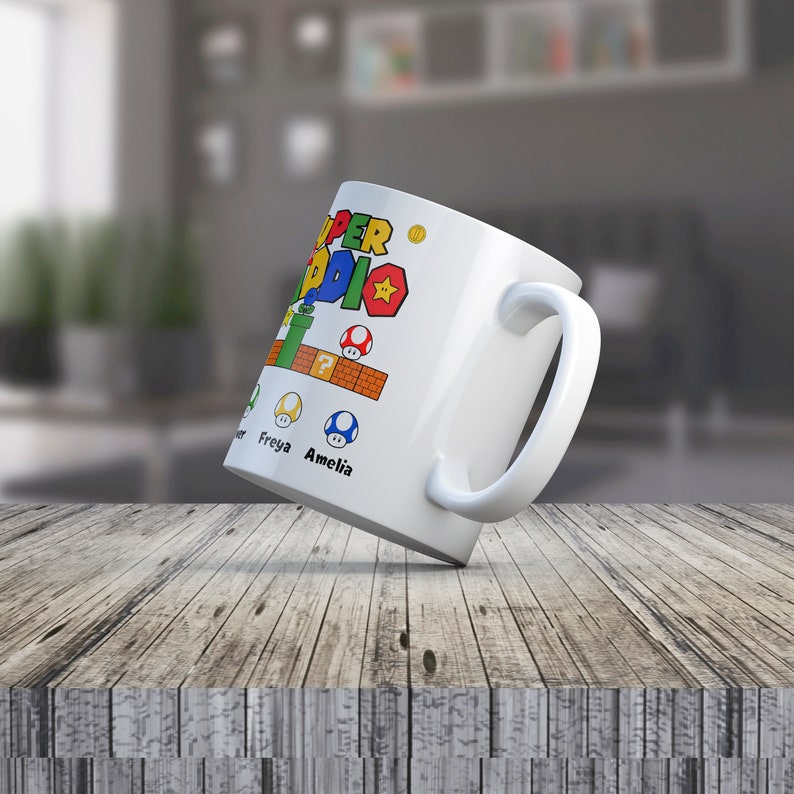 May include: White ceramic mug with a colorful Super Mario Bros. design. The mug features the text "Super Mario" and a yellow star. There are also images of a green pipe, a question mark block, and three mushrooms. The text "Freya" and "Amelia" are written below the mushrooms.