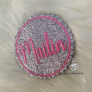 Könnte beinhalten: Ein silberner Glitzerkreis mit einem rosa gestickten Namen "Mailin" in einer kursivschrift.