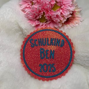 Könnte beinhalten: Ein roter, glitzernder runder Aufnäher mit dem Text "SCHULKIND BEN 2025" in Türkis. Der Aufnäher hat einen türkisfarbenen Rand und einen gewellten Rand. Rosa Blumen sind im Hintergrund.