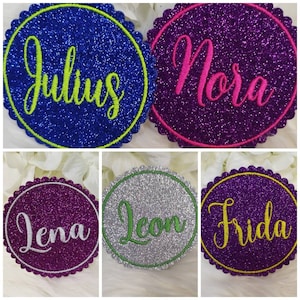 Könnte beinhalten: Fünf runde, glitzernde, farbige, personalisierte Namensschilder mit gewellten Rändern. Die Namen sind Julius, Nora, Lena, Leon und Frida.
