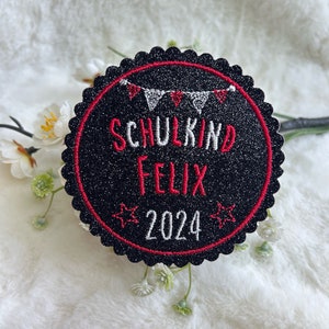 Könnte beinhalten: Runder schwarzer und roter Patch mit dem Text "Schulkind Felix 2024" und einer Girlande aus roten und weißen Flaggen.