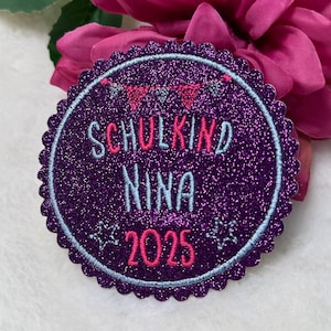 Könnte beinhalten: Ein runder, lila Glitzeraufnäher mit dem Text "SCHULKIND NINA 2025" in Rosa und Hellblau. Der Aufnäher hat einen gewellten Rand und einen hellblauen Rand. Kleine Sterne und ein Banner-Design sind ebenfalls enthalten.