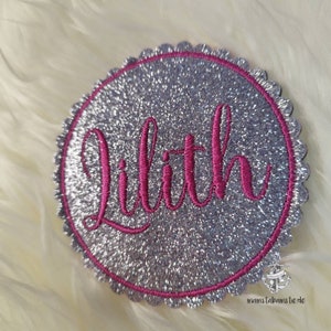 Könnte beinhalten: Ein silberner Glitzerkreis mit einem rosa gestickten Rand und dem Namen "Lilith" in rosa Schrift.