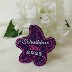 Könnte beinhalten: Lilafarbener, glitzernder Stern-Patch mit rosa Nähten. Der Patch trägt die Aufschrift "Schulkind Tilda 2023" in weißer Stickerei.