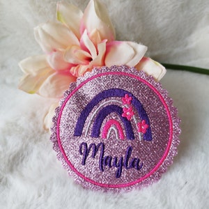 Könnte beinhalten: Rosa glitzernder Bügelaufkleber mit einem violetten Regenbogen und drei rosa Blumen. Der Aufnäher ist mit "Mayla" in violetter Stickerei versehen.