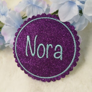 Könnte beinhalten: Ein runder, lila Glitzeraufnäher mit einem gewellten Rand und einem hellblauen Rand. Der Name "Nora" ist in hellblauer Schrift über die Mitte gestickt. Der Aufnäher ist wahrscheinlich für Kleidung oder Accessoires.
