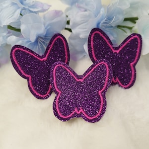 Könnte beinhalten: Drei violette Glitzer-Schmetterlingsapplikationen mit rosa Umrandungen. Die Schmetterlinge befinden sich auf einer weißen, flauschigen Oberfläche.