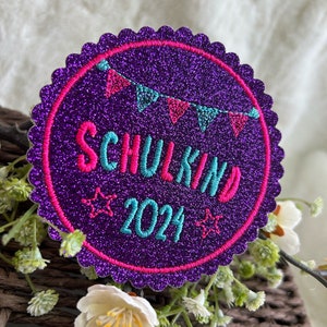 Könnte beinhalten: Lilafarbener Glitzer-Kreis-Patch mit rosa und blauer Stickerei. Der Patch sagt "Schulkind 2024" mit Sternen auf beiden Seiten.