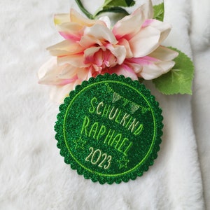 Könnte beinhalten: Grüner Glitzerkreis mit dem Text "Schulkind Raphael 2023" in weißer Stickerei.