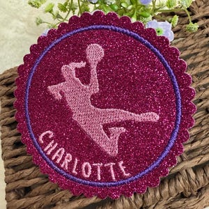 Könnte beinhalten: Ein runder Glitzer-Patch in Pink mit violetter Umrandung und gewellten Kanten. Der Patch zeigt eine rosa Silhouette eines Basketballspielers mit dem Namen "CHARLOTTE" in Violett darunter.