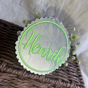 Könnte beinhalten: Ein runder, silberfarbener Namensschild mit einem grünen gewellten Rand und einem grünen Kreis um den Namen "Henrik", der in grün gestickt ist.