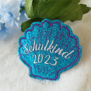 Könnte beinhalten: Eine blaue und lila glitzernde Muschel mit dem gestickten Text "Schulkid 2023" in weiß.