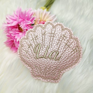 Könnte beinhalten: Eine rosa Stoffmuschel mit dem goldenen Sticknamen "Lila" darauf.