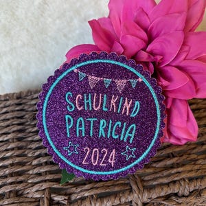Könnte beinhalten: Lilafarbener Glitzer-Kreis-Patch mit türkisfarbenem Rand und weißem Text "Schulkind Patricia 2024" mit rosa und weißen Flaggen über dem Text.