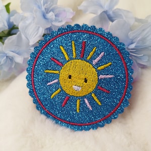 Könnte beinhalten: Ein blauer und roter gestickter Aufnäher mit einer lächelnden gelben Sonne mit rosa und roten Strahlen.
