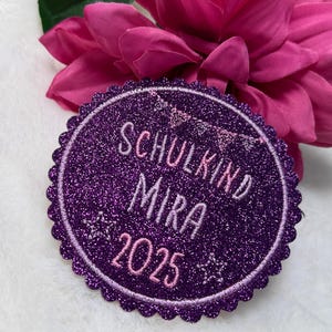 Könnte beinhalten: Ein runder, glitzernder, violetter Aufnäher mit einem gewellten Rand. Der Aufnäher zeigt die Worte "SCHULKIND MIRA 2025" in hellrosa Schrift. Dekorative Sterne und ein Banner sind ebenfalls im Design enthalten. Der Aufnäher befindet sich vor dem Hintergrund einer rosa Blume.