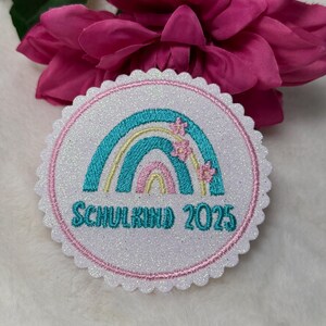 Könnte beinhalten: Ein runder, weißer Aufnäher mit einem gewellten Rand, der ein Regenbogen-Design in Türkis, Gelb und Rosa mit kleinen rosa Blütenakzenten zeigt. Der Text "SCHULKIND 2025" ist türkis gestickt. Der Aufnäher hat ein glitzerndes Aussehen.