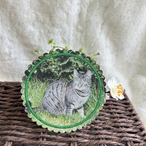 Könnte beinhalten: Ein runder Untersetzer mit gewelltem Rand, der ein gedrucktes Bild einer grauen getigerten Katze im grünen Gras zeigt. Der Untersetzer hat einen grünen, gestickten Rand um das Katzenbild, das vor einem natürlichen Hintergrund platziert ist.