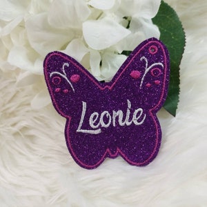 Könnte beinhalten: Lilafarbener Glitzer-Schmetterling mit rosa Akzenten und dem Namen Leonie in weiß gestickt.