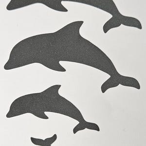 Könnte beinhalten: Vier dunkelgraue Delfin-Silhouetten unterschiedlicher Größe sind vertikal vor einem weißen Hintergrund angeordnet. Die Delfine haben ein strukturiertes Aussehen, das auf ein glitzerndes oder körniges Material hindeutet. Das Bild ist einfach und grafisch.