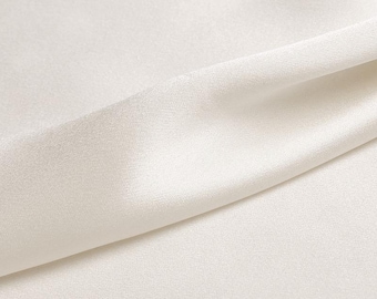 May include: Close-up of a cream-colored fabric with a subtle sheen. The fabric is draped, creating soft folds and shadows. The texture appears smooth and luxurious, suggesting a high-quality material.