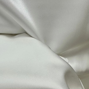May include: A close-up of a white satin fabric, showcasing its smooth and silky texture. The fabric is folded to highlight its drape and sheen.
