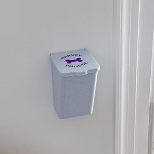 May include: A light blue pet treat container mounted on a white wall. The lid features the names "HARVEY" and "PHOEBE" in purple, along with a bone graphic. The container is rectangular with a slightly textured surface.