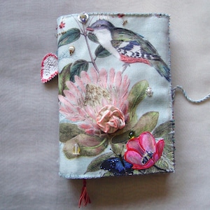 Könnte beinhalten: Ein stoffbezogenes Notizbuch mit Blumen- und Vogelmuster. Das Cover zeigt einen Vogel, der auf einem Ast über einer großen rosa Blume und einer roten Tulpe sitzt. Verzierungen sind Perlen und eine Quaste. Das Notizbuch wird mit einer Schnur verschlossen.