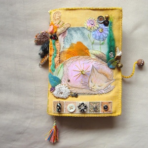 Puede incluir: Un diario cubierto de tela amarilla con un gato durmiente bordado, motivos florales y varios botones decorativos. La cubierta incluye una borla multicolor en la parte inferior, un cordón amarillo con una cuenta de madera y adornos de tela.