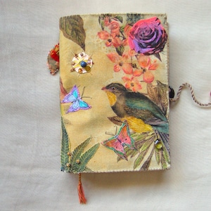 Op de afbeelding: Een rechthoekig, met stof bekleed notitieboek met een gele achtergrond en een print van een vogel, vlinders, een grote roos en bladeren. De kaft is versierd met kleine metalen studs, kralen en een koord met een rode kraal.