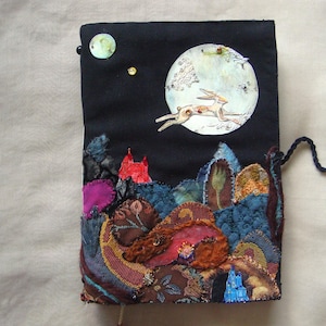 May include: A rectangular fabric journal cover featuring a black background with a large white moon and a leaping hare. The lower section displays layered, textured fabric patches in shades of blue, red, brown, and magenta, with a cord tie closure.