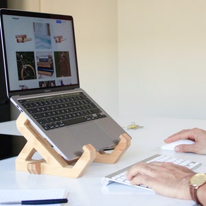 Może przedstawiać: Laptop umieszczony na drewnianym stojaku do laptopów. Laptop jest otwarty i wyświetla stronę internetową ze zdjęciami drewnianych stojaków do laptopów. Osoba używa bezprzewodowej klawiatury i myszy.